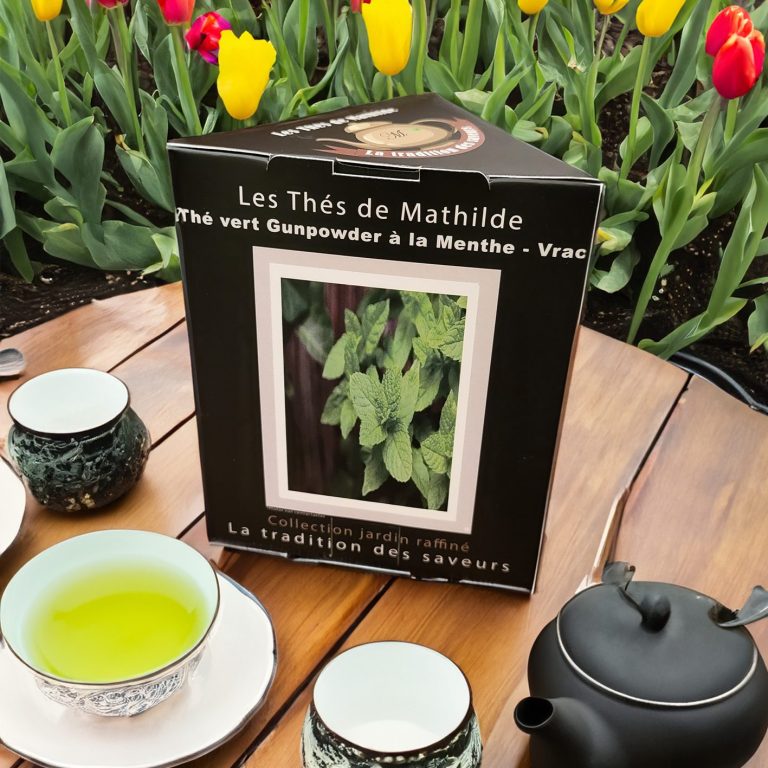 Les Thés de Mathilde - Thés vert à la Menthe Boîte de thé à la menthe avec des tasses, une théière et des tulipes colorées en arrière-plan.